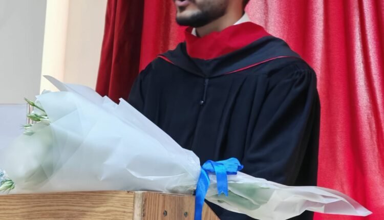 Male graduate in a black gown and red sash speaks at a wooden podium, with a white bouquet nearby, red stage curtain behind.