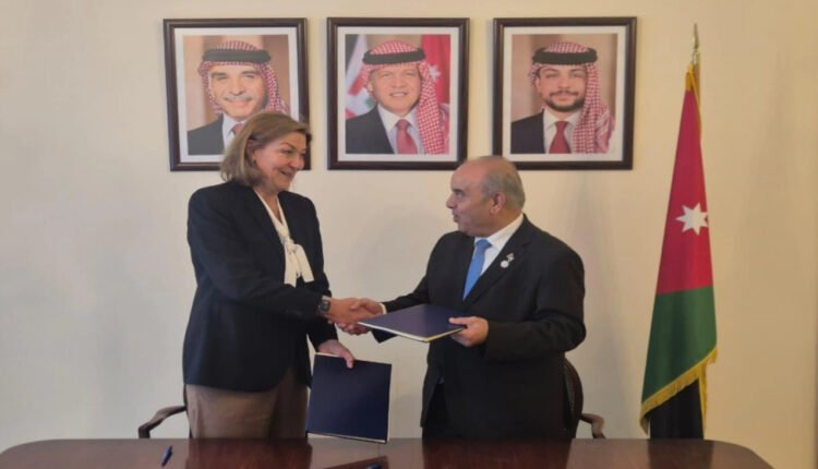Two professionals shake hands across a table during a signing ceremony, with Jordan’s flag in the background and portraits on the wall nearby.