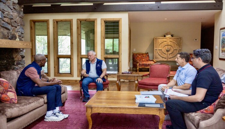 Four men sit in a cozy living room around a wooden coffee table, engaged in a discussion; one man speaks while others listen.