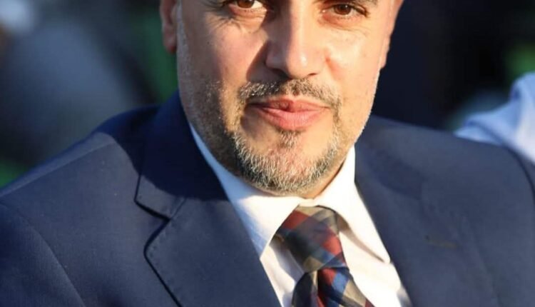 Head-and-shoulders portrait of a middle-aged man in a navy suit and plaid tie outdoors, looking at the camera with a slight smile.