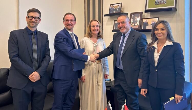 Five professionals in suits in an office, two men shake hands while others smile; German and Jordanian flags on the table nearby.